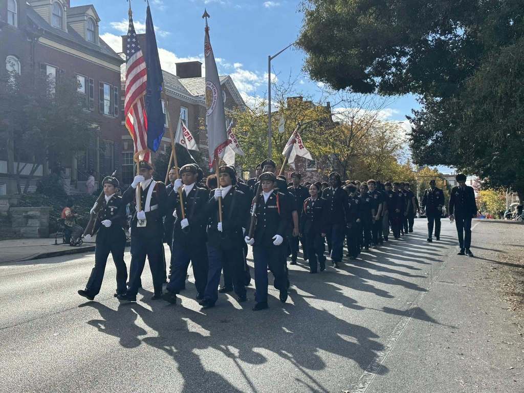 A group of uniformed individuals are marching down a street outside holding flags, including the American flag.