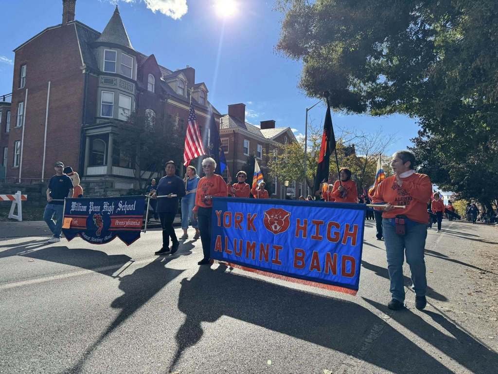 A parade featuring the William Penn Senior High School Bearcat Marching Band and the York High Alumni Band. A group of individuals are carrying banners. Some people are holding flags.