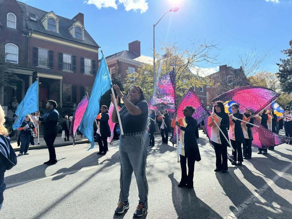 A lively parade scene with performers waving vibrant blue and pink flags under a sunny sky, showcasing community spirit and celebration.