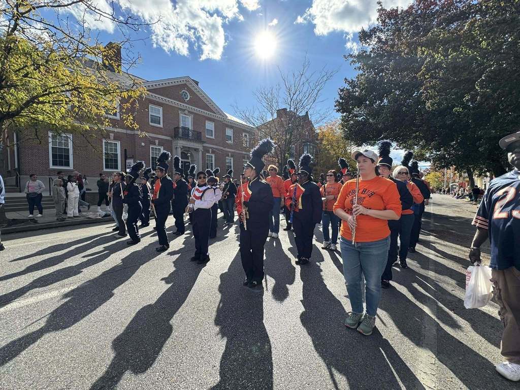 A lively parade scene with musicians in uniforms, vibrant costumes, and spectators under a sunny sky and autumn foliage.