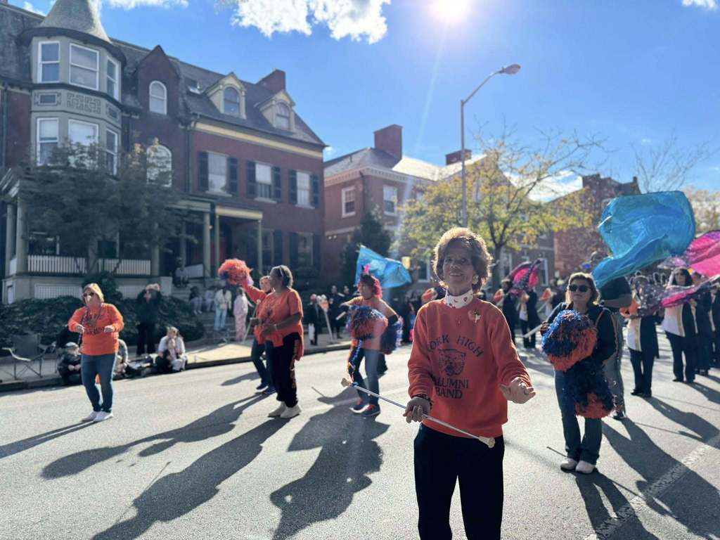 A vibrant parade scene with participants in bright orange shirts, pom-poms, and colorful flags, set against a sunny backdrop.