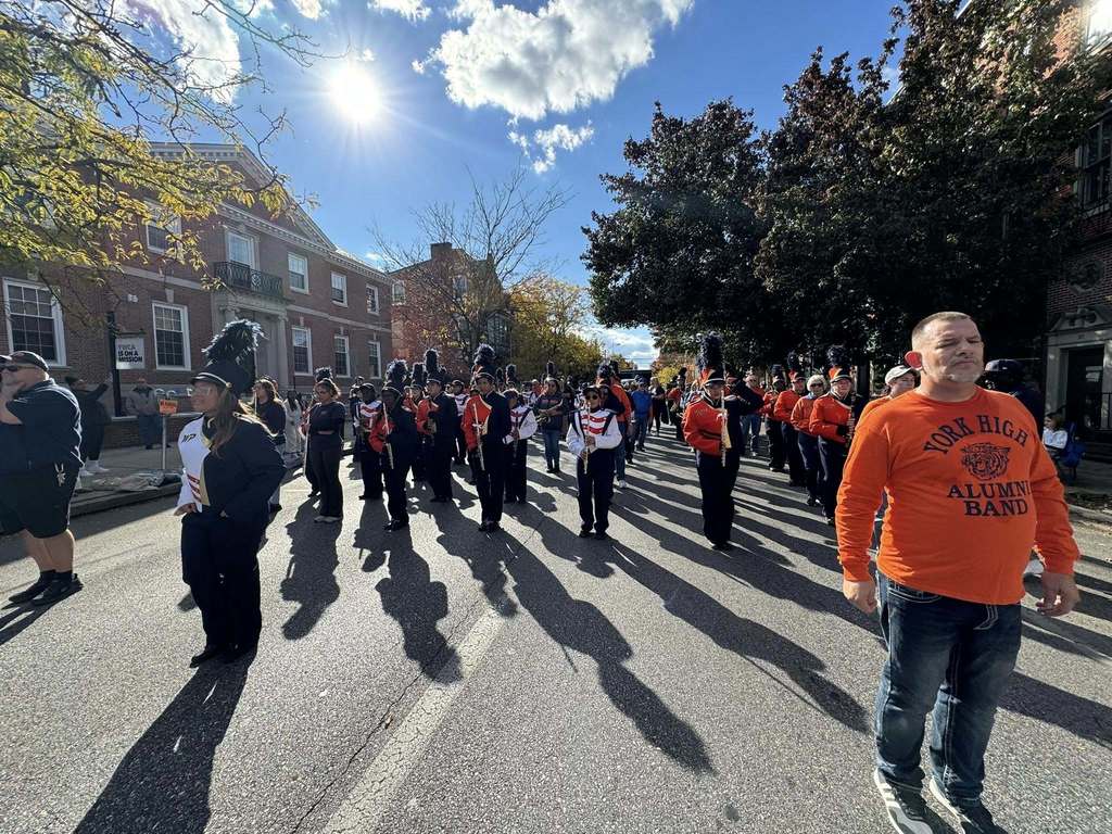 A marching band is parading down a sunny street, with members in dark uniforms and feathered hats. Vibrant fall foliage and historic brick buildings line the street.