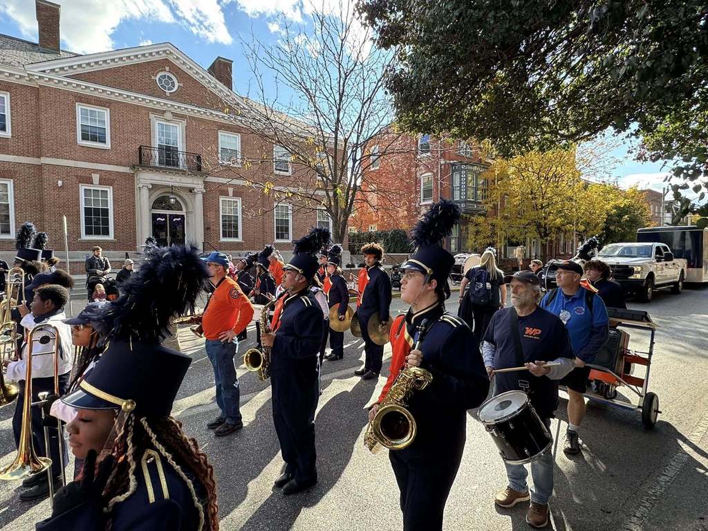 A marching band in uniforms prepares for a performance on a street lined with trees, with a large brick building in the background.