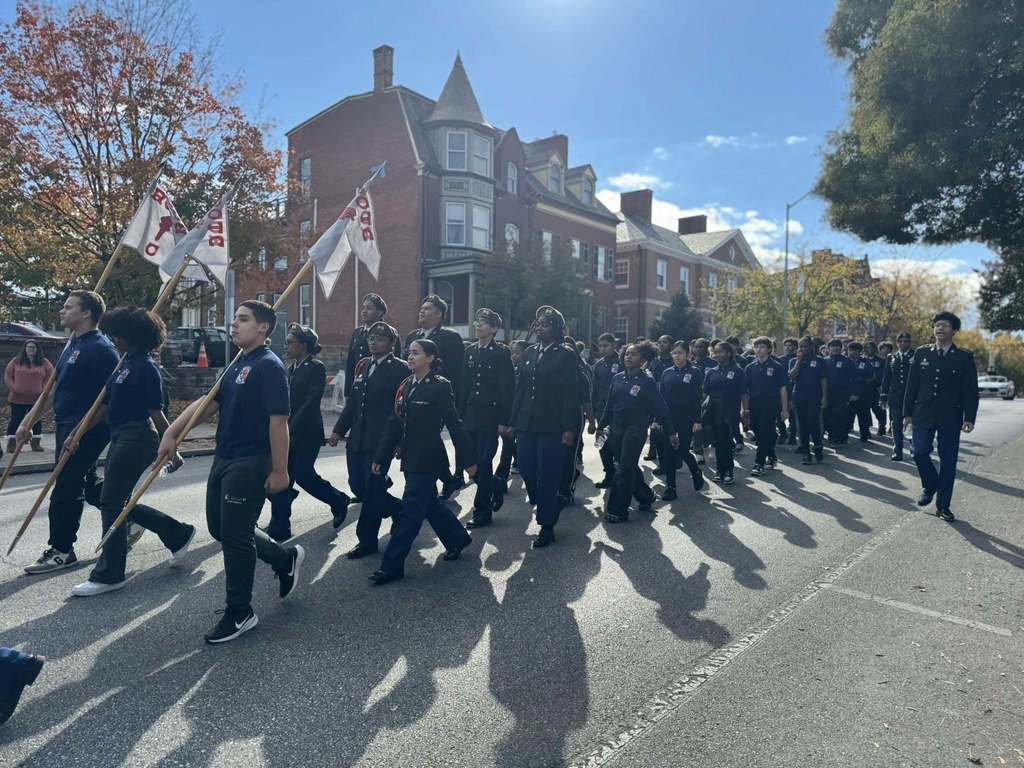 A large group of people wearing uniforms are marching in formation outside along a street. Some of the people are carrying flags.