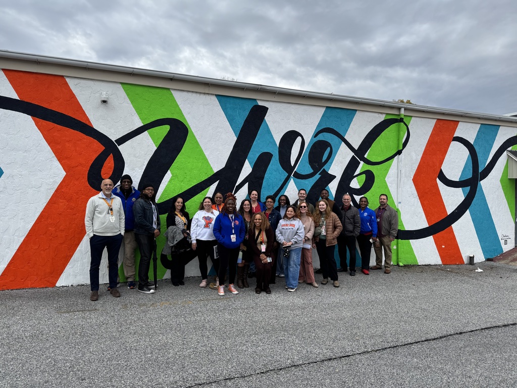 A group of district staff members are standing in front of a colorful mural with bold diagonal stripes in orange, green, and blue, overlayed with black script writing.