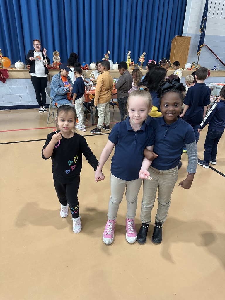 A group of students are holding hands in a school gym that is decorated for an event, with pumpkins and autumn-themed decor in the background. Other students and adults can also be seen in the background.