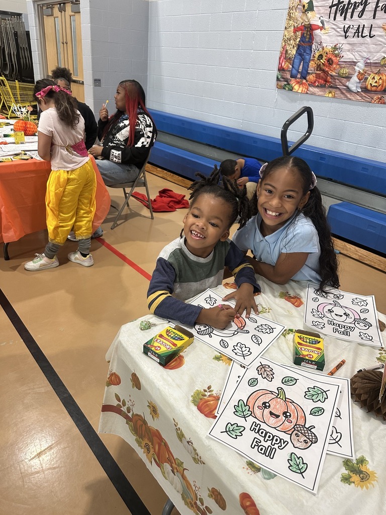 Two children are sitting at a table with autumn decorations, coloring fall-themed worksheets in a school gym. Other students and adults can be seen in the background.