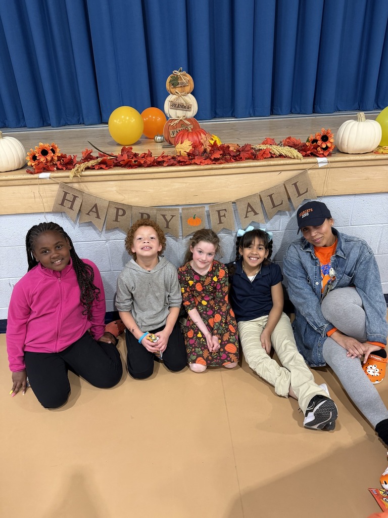 A group of students and a district staff member are sitting in front of a "Happy Fall" banner, surrounded by autumn decorations, pumpkins, and colorful balloons.