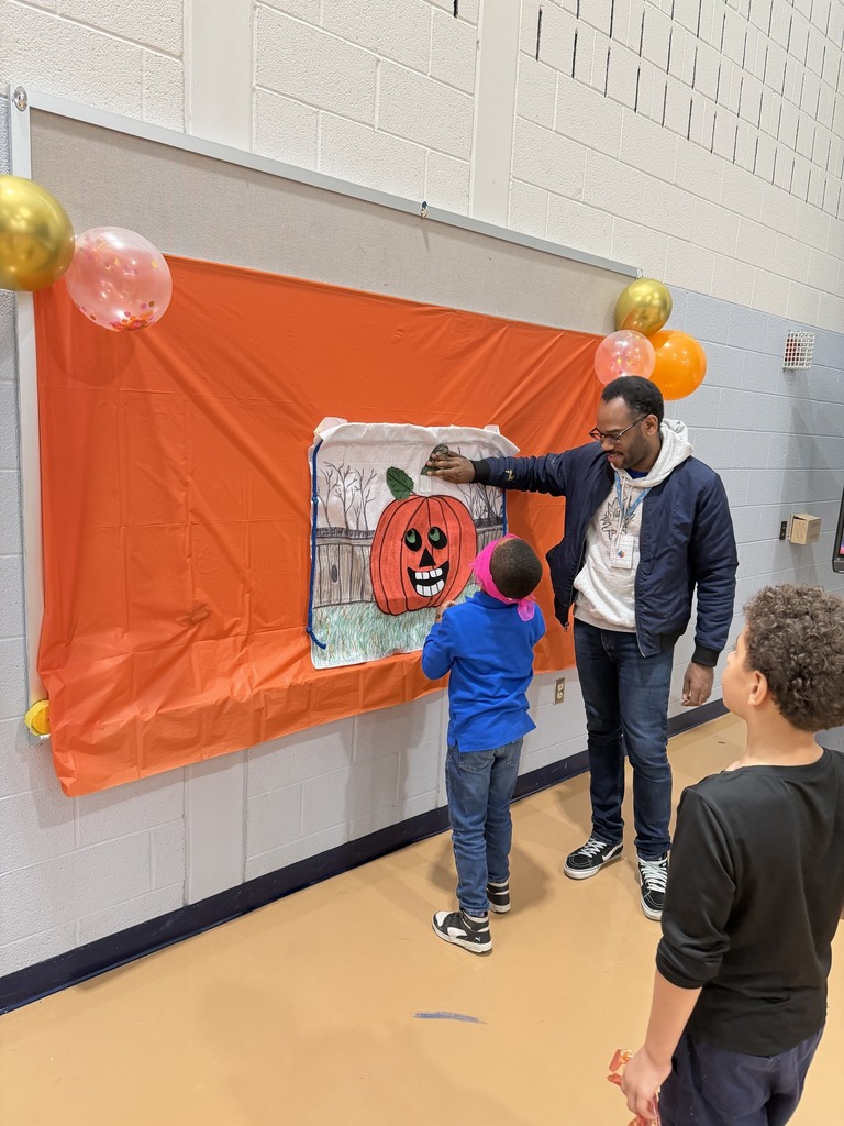 Two students are engaging in a festive activity in a school gym at a decorated wall featuring a large, colorful pumpkin drawing with balloons nearby. An instructor is helping the students.