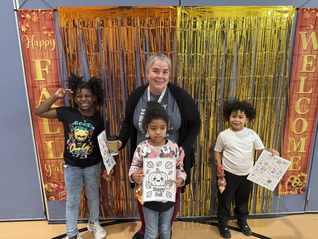A district staff member and three students are posing in front of a festive gold and orange tinsel backdrop that says "Happy Fall Y'all" and "Welcome" on the sides. The district staff member and two of the students are holding up fall-themed coloring pages and snacks. 