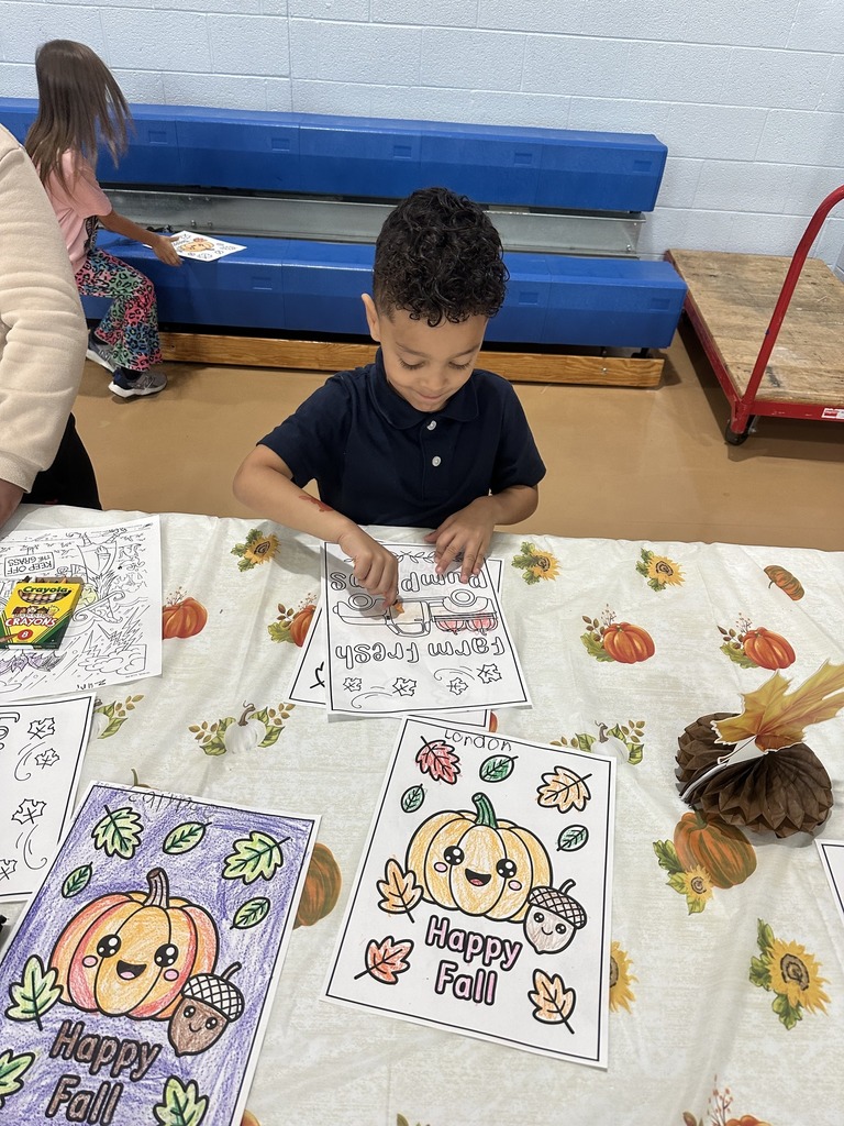 A student is sitting at a table with autumn decorations, coloring fall-themed drawings, surrounded by crayons. Another student can be seen in the background.