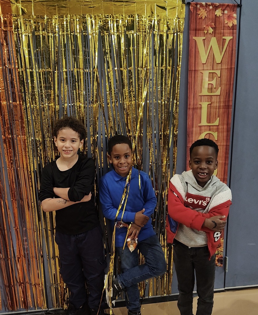 Three students are standing in front of a gold and orange tinsel backdrop with a “Welcome” banner on the side. They are posing with their arms crossed. 