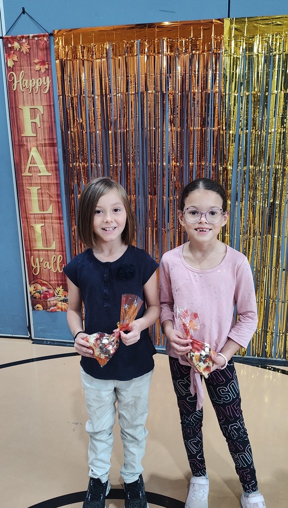 Two students are holding treat bags while standing in front of a festive autumn backdrop that says "Happy Fall Y'all" in a school gym.