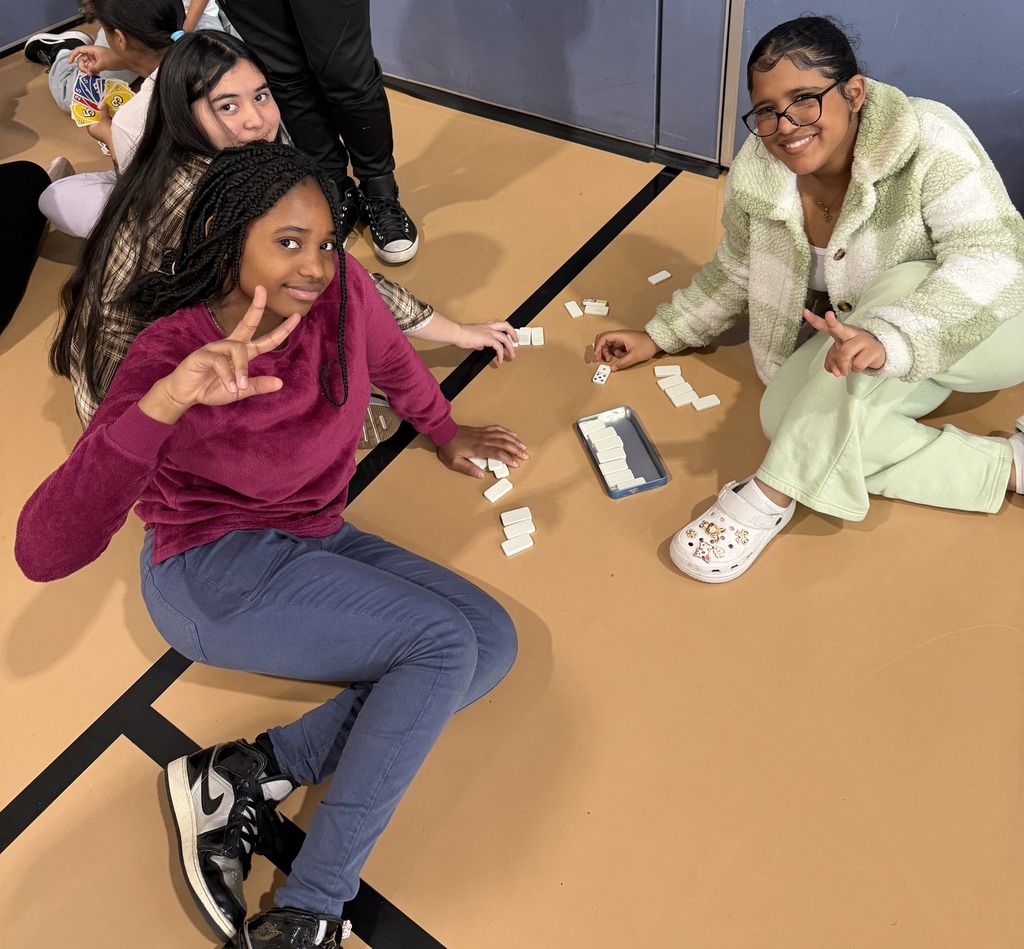 Three students are sitting on a gym floor playing a game with white dominoes and dice. Another student can be seen in the background.