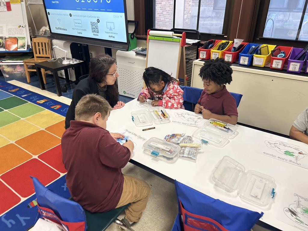 A group of three students and an adult are sitting at a table in a classroom. The students are working on an activity and the adult is observing them.