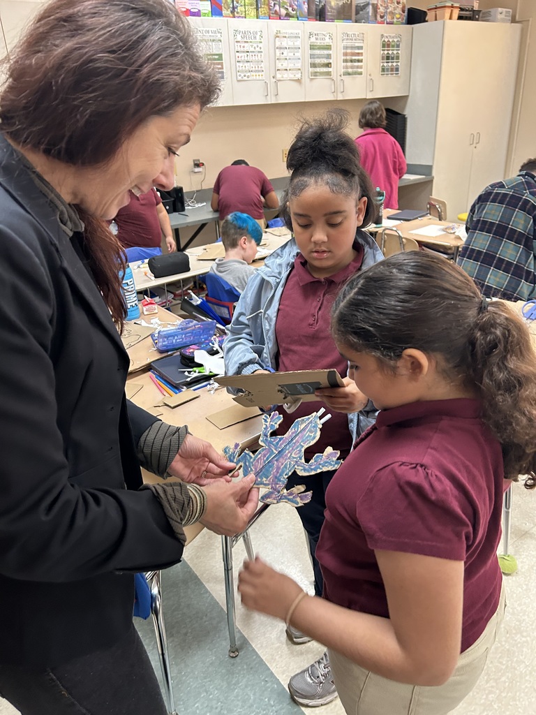 An adult is observing a craft in her hand while two students are standing nearby in a classroom. Other students can be seen in the background.