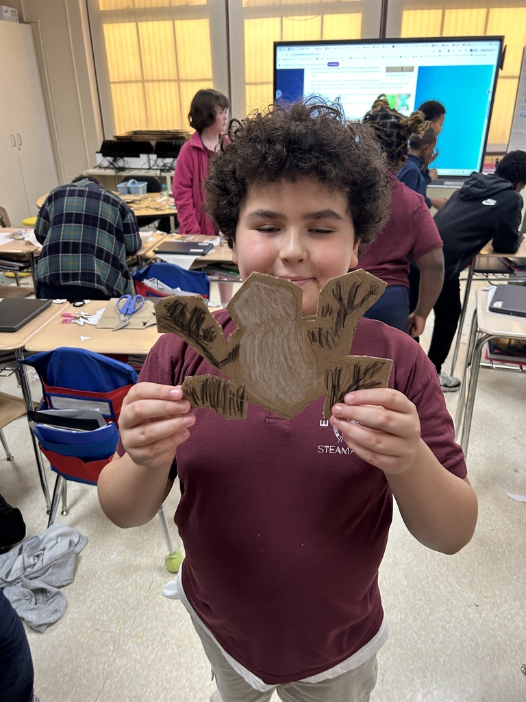 A student is holding up a craft in his hand while other students are in the background in a classroom.
