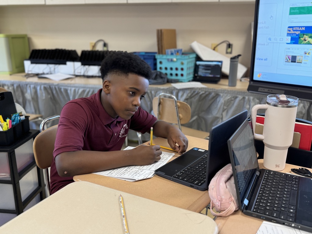A student sitting at a desk in a classroom is writing on a worksheet while looking at a laptop. 