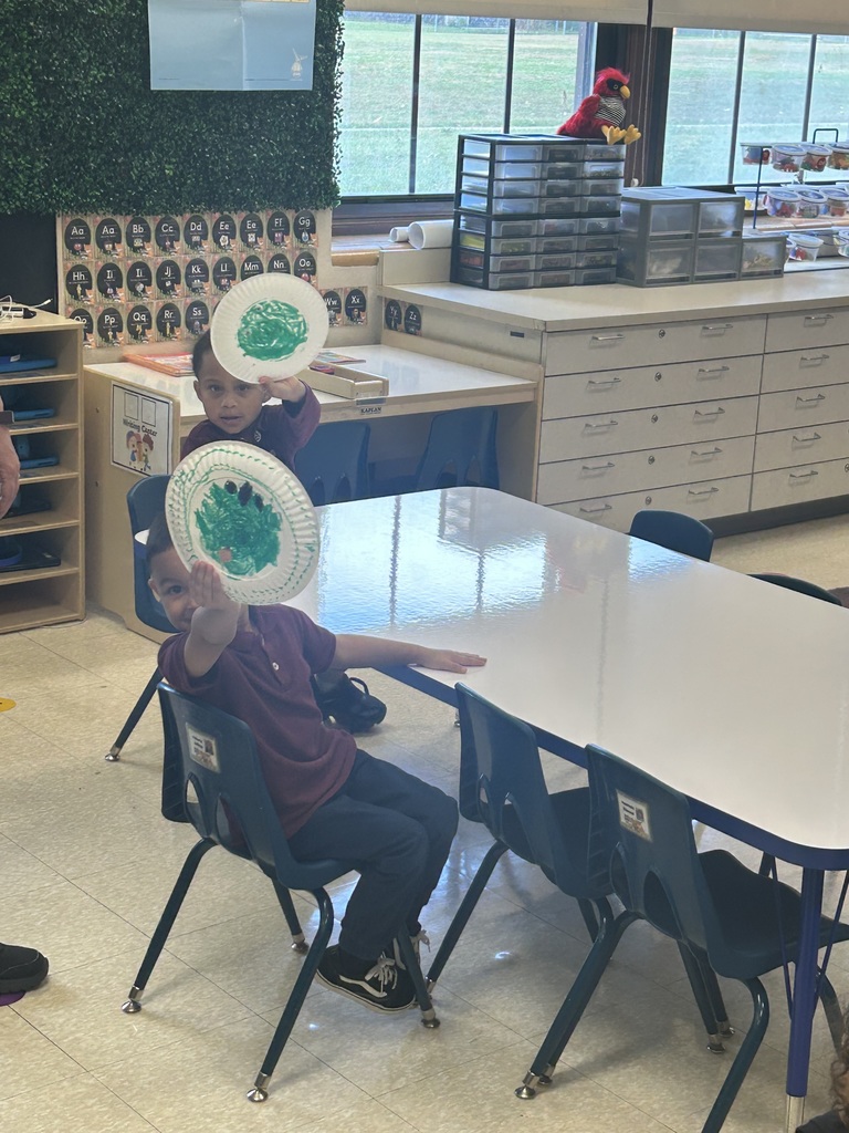 Two students sitting at a table in a classroom are holding up paper plates with green and black paint on it. 