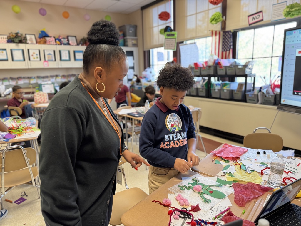 A district staff member and a student are working together on a colorful art project in a classroom. Other students can be seen in the background.