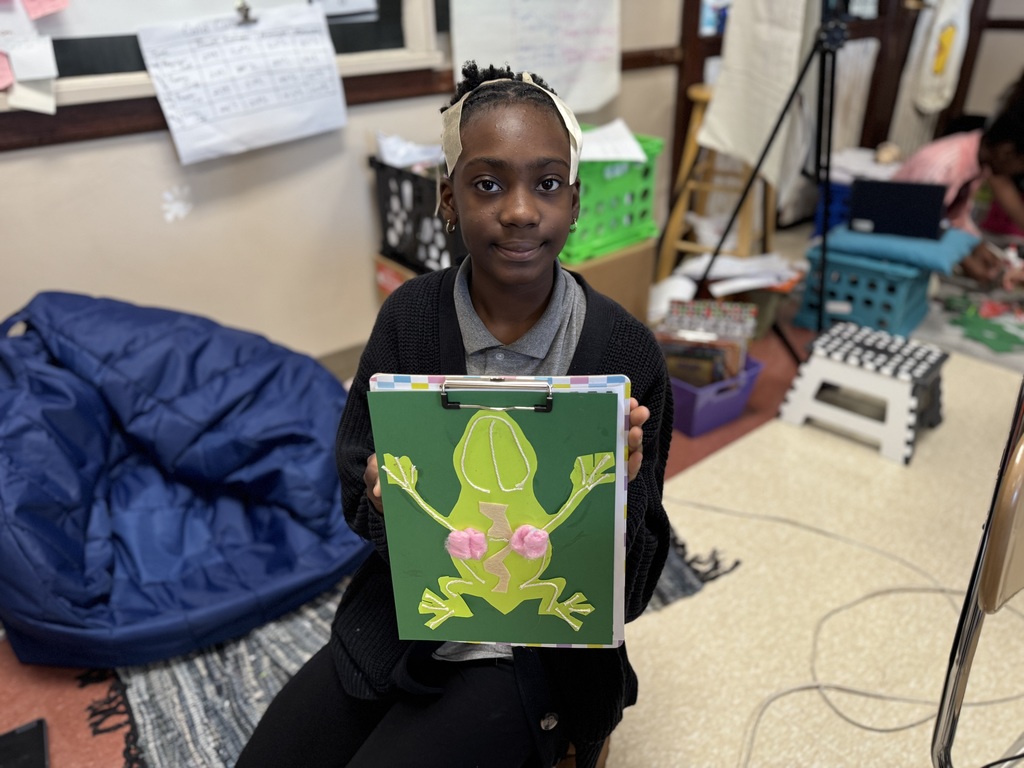 A student is sitting in a classroom holding up a green art project featuring a cutout of a frog with pink cotton balls attached. The background includes posters, bins, and a bean bag chair in the background.
