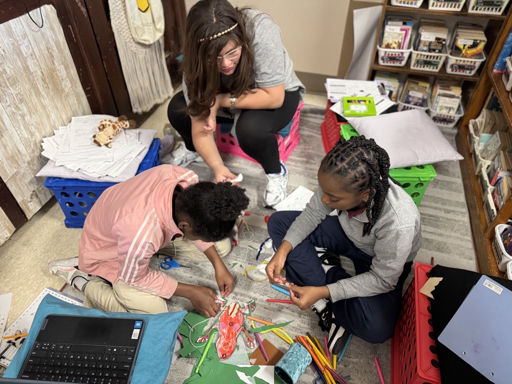 Three students are sitting on the floor in a classroom, engaging in crafts with colored papers and markers. They are surrounded by papers and supplies.