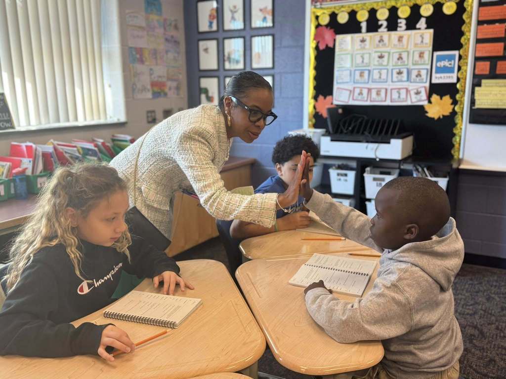 A district staff member is giving a high five to a student who is sitting at a desk in a classroom. Two other students are sitting at desks nearby.