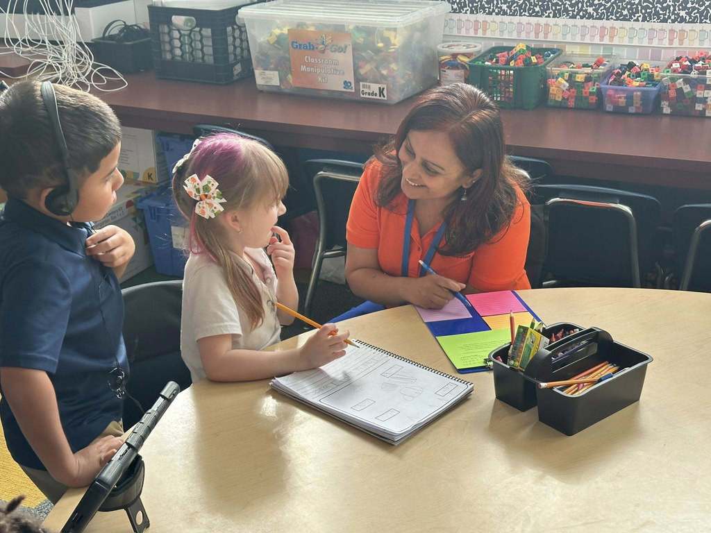 A district staff member is sitting at a table in a classroom observing two other students with a pencil and clipboard.
