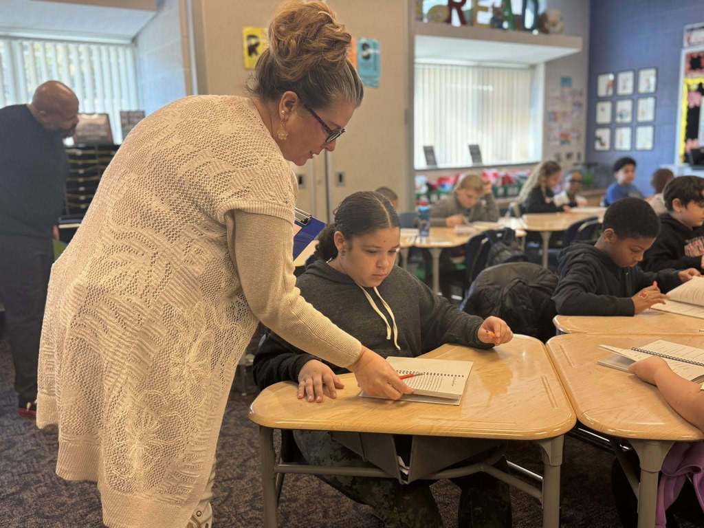 A district staff member is standing over a student in a classroom observing her with a pen. Another district staff member and other students can be seen in the background.