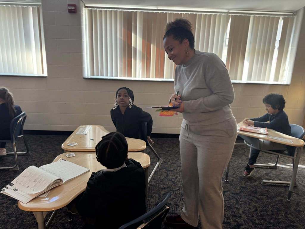 A district staff member is standing over a student in a classroom observing him with a pen and clipboard. Other students can be seen in the background.