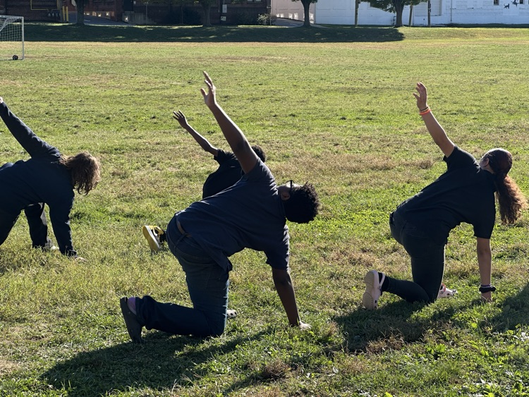 A group of students kneeling down on grass with their left arms lifted in the air.