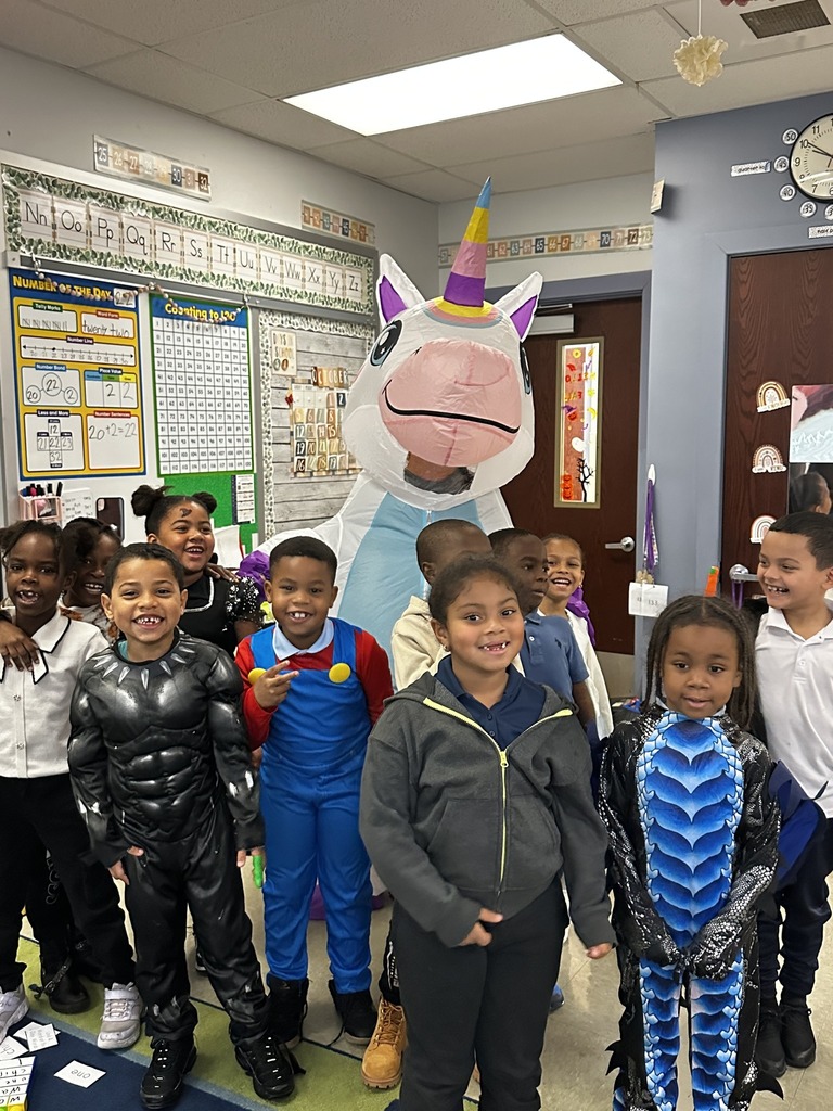 A colorful classroom scene featuring children in costumes with a large unicorn mascot, surrounded by educational materials and decorations.