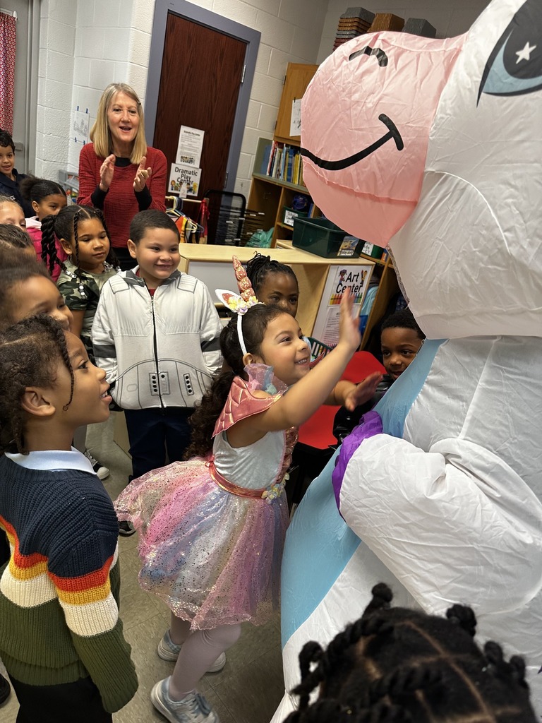 Children in colorful costumes excitedly interacting with a large inflatable unicorn in a classroom setting.