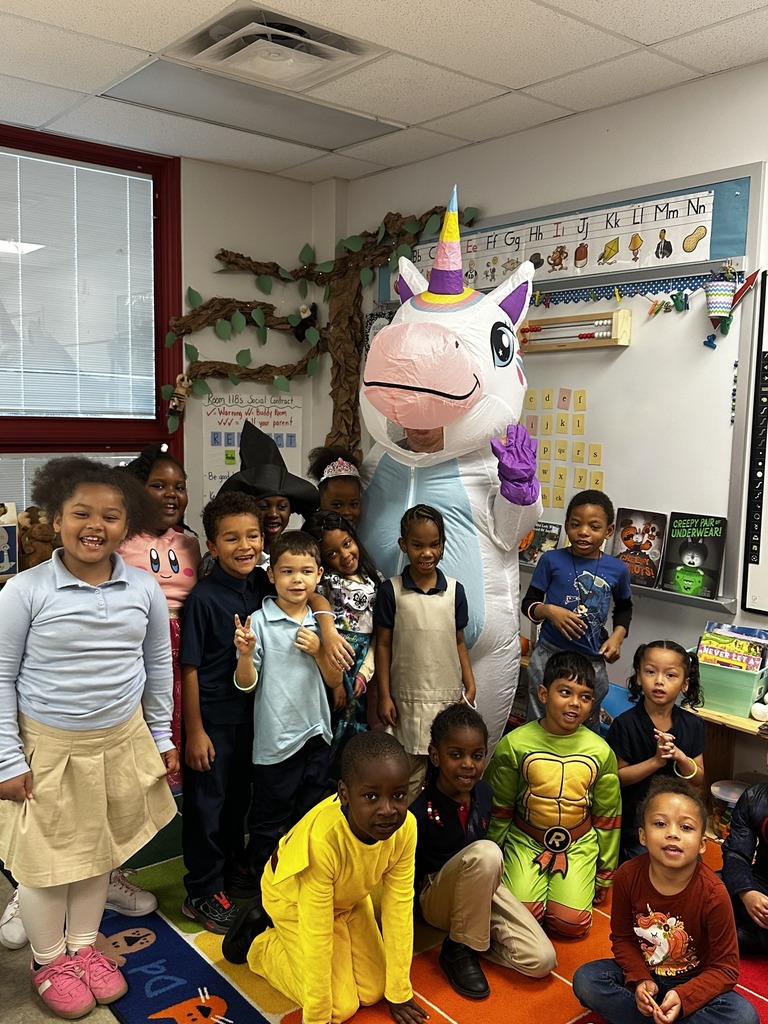 A colorful classroom scene featuring children in costumes with a large unicorn mascot, surrounded by educational materials and decorations.
