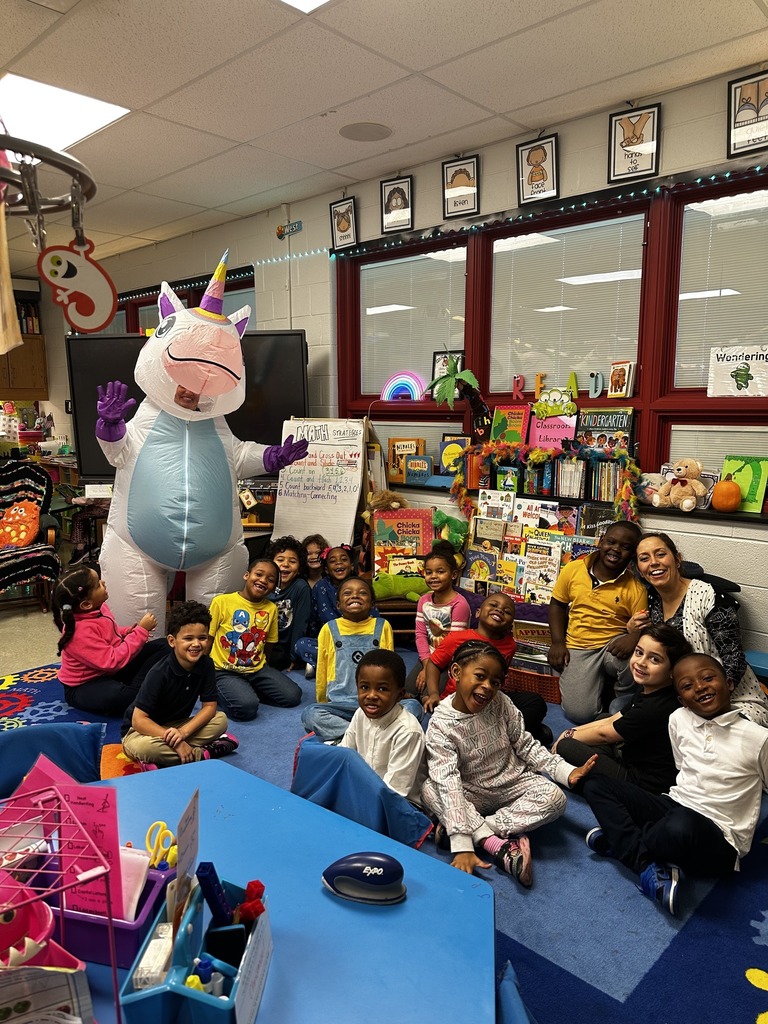 A colorful classroom scene featuring children in costumes with a large unicorn mascot, surrounded by educational materials and decorations.