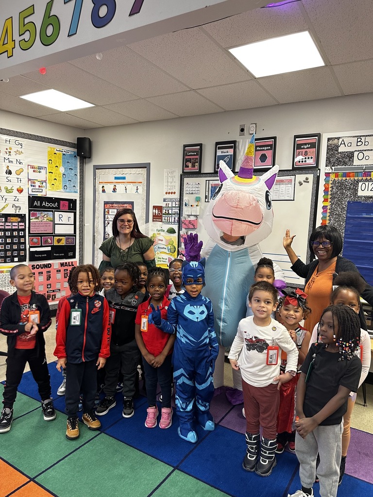A colorful classroom scene featuring children in costumes with a large unicorn mascot, surrounded by educational materials and decorations.