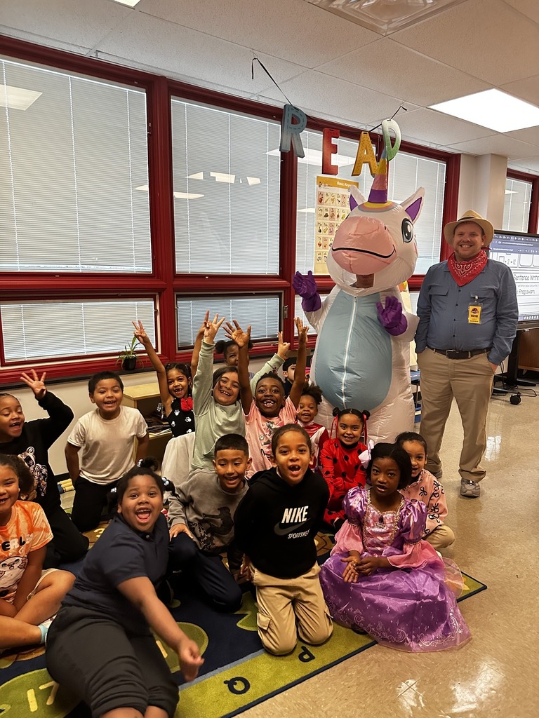 A colorful classroom scene featuring children in costumes with a large unicorn mascot, surrounded by educational materials and decorations.