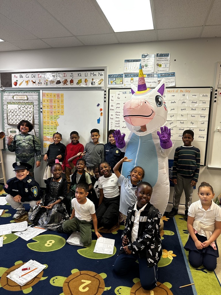 A colorful classroom scene featuring children in costumes with a large unicorn mascot, surrounded by educational materials and decorations.