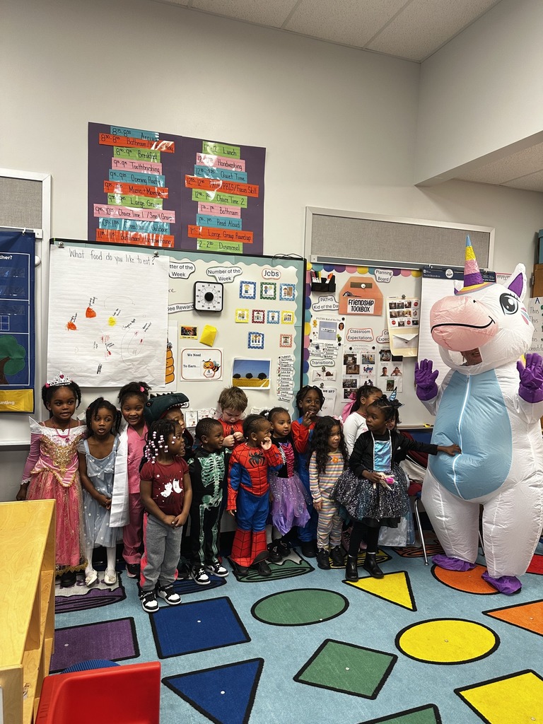 A colorful classroom scene featuring children in costumes with a large unicorn mascot, surrounded by educational materials and decorations.