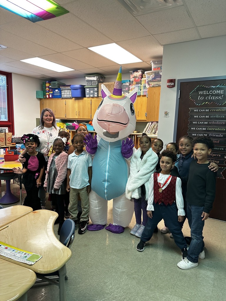 A colorful classroom scene featuring children in costumes with a large unicorn mascot, surrounded by educational materials and decorations.