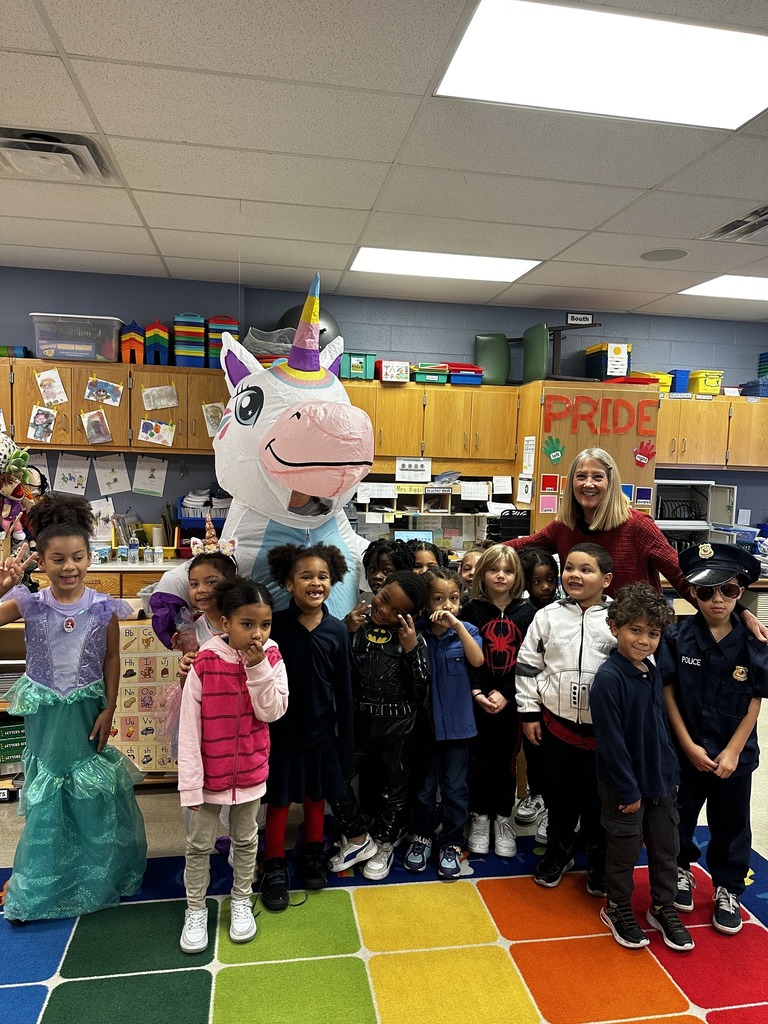 A colorful classroom scene featuring children in costumes with a large unicorn mascot, surrounded by educational materials and decorations.