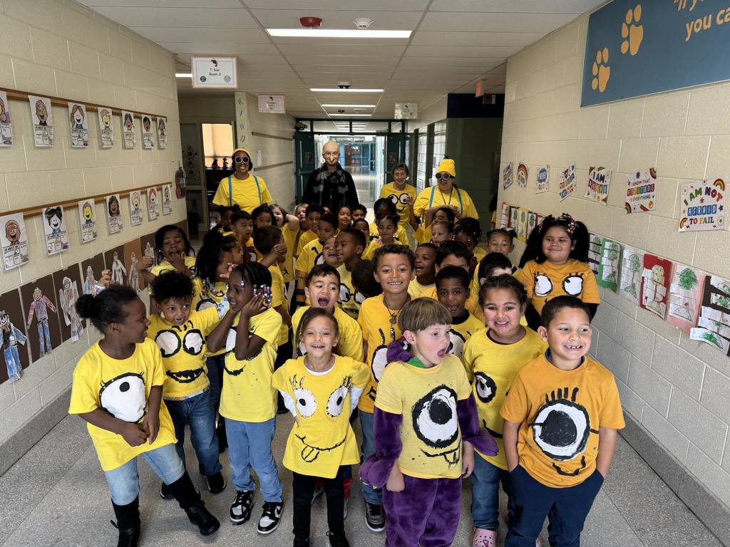 A group of students and four adults dressed in minion-themed shirts standing in a school hallway adorned with colorful artwork and educational posters.