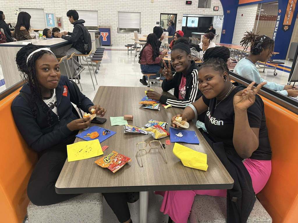 A group of students enjoying snacks at a school cafeteria table, with various colorful wrappers and notes scattered around. Other people can be seen in the background.