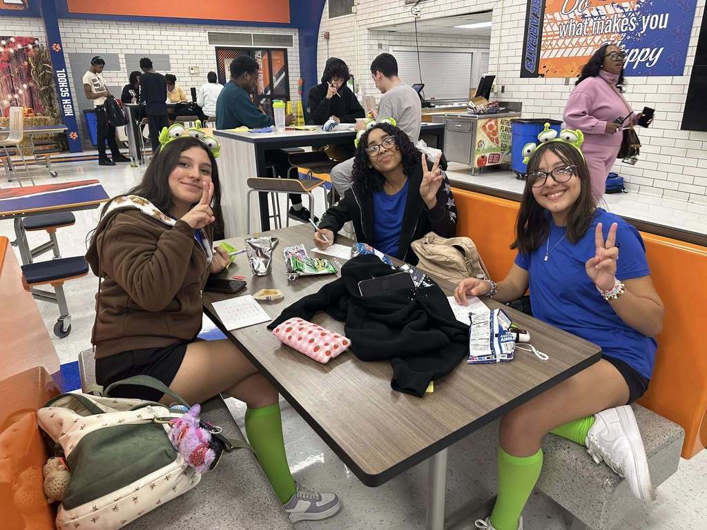 A group of students in blue shirts and green socks are holding up the peace sign while sitting at a table in a school cafeteria. Backdrop features school decorations. Other people can be seen in the background.