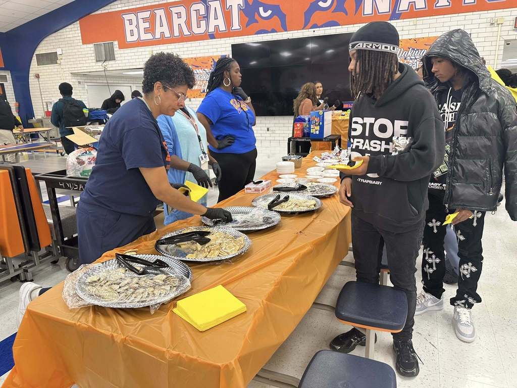 A school cafeteria event featuring a table with food trays, district staff members serving students, and festive decorations visible in the background. Other people can be seen in the background.