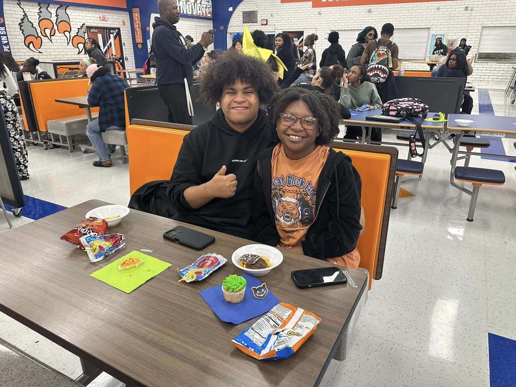 Two students sitting at a dining table in a school cafeteria with snacks, cupcakes. The background shows various people socializing. 