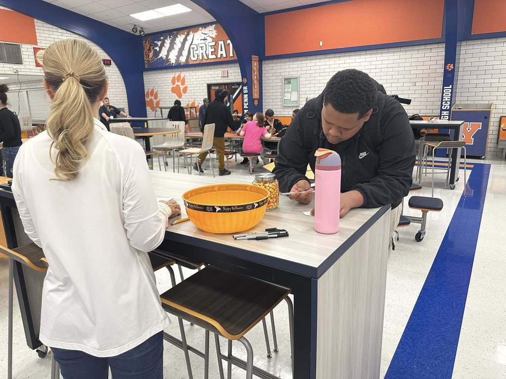 A district staff member is standing near a table in a school cafeteria, watching as a student has a pen in his hand. A jar of candy corn, a pink water bottle and multiple pens are on the table. Other people can be seen in the background.