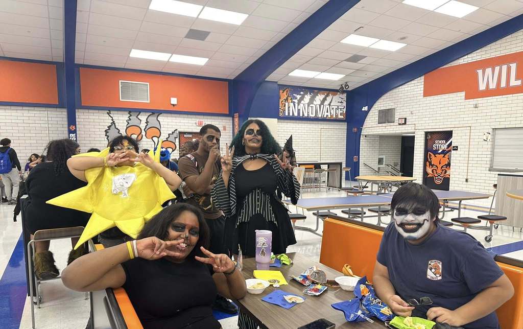 A group of students gathering around a table in a school cafeteria, some dressed in costumes, enjoying snacks and posing playfully. Other people can be seen in the background.