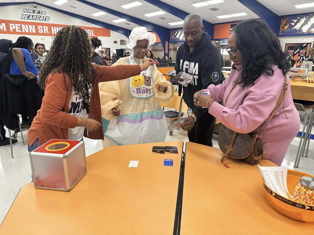 District staff members and a student are gathered around an orange table in a school cafeteria. The student is wearing a humorous baby costume, and district staff members are standing nearby. Other people can be seen in the background.