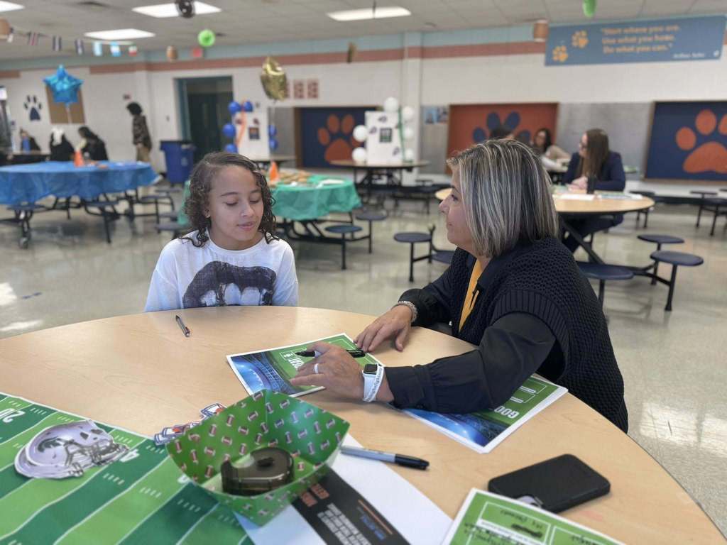 A district staff member and a young student are sitting at a round table in a school cafeteria, engaging in a discussion. Other people can be seen in the background.